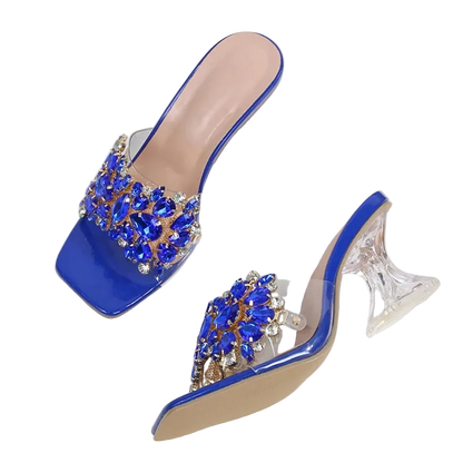 A pair of women's mule high heels with glossy blue patent leather uppers adorned with large blue and clear rhinestone embellishments, clear block heels, and beige insoles, displayed on a white fabric surface.