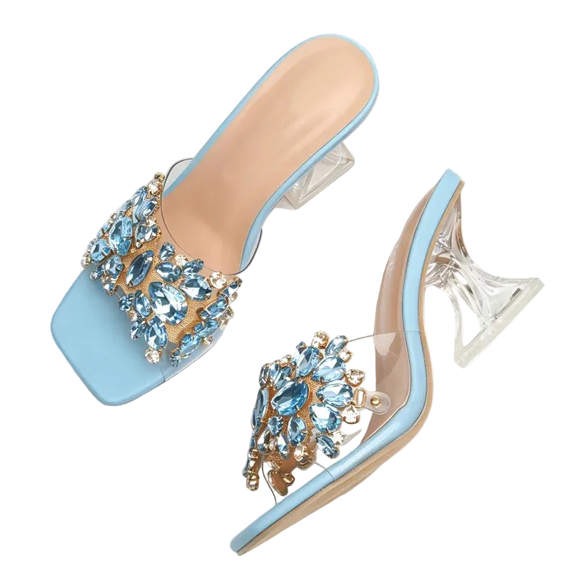 A pair of light blue square-toe mule sandals with clear high heels and ornate crystal embellishments on the straps, displayed on a white fabric surface with a denim and woven strap prop.