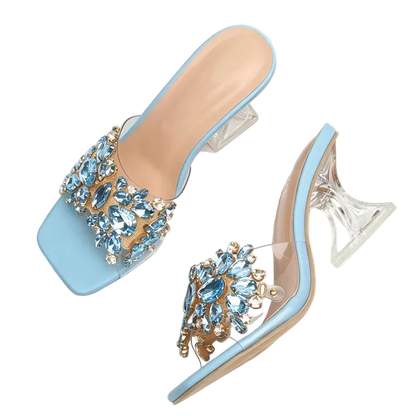A pair of light blue square-toe mule sandals with clear high heels and ornate crystal embellishments on the straps, displayed on a white fabric surface with a denim and woven strap prop.
