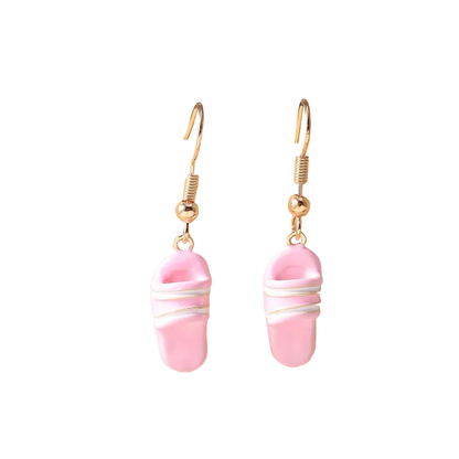 A pair of gold-tone dangle earrings with pink slipper-shaped charms wrapped in white wire, displayed on a beige ceramic stand.