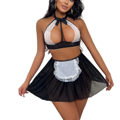 A woman models a black and white lingerie set featuring a lace halter bra, a sheer black mini skirt, and a white apron with a black bow, set in a bedroom environment.